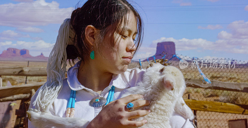 在一个阳光明媚的日子里，亚利桑那州/犹他州的纪念碑谷，一个十几岁的美国土著女孩把一只羔羊抱在怀里